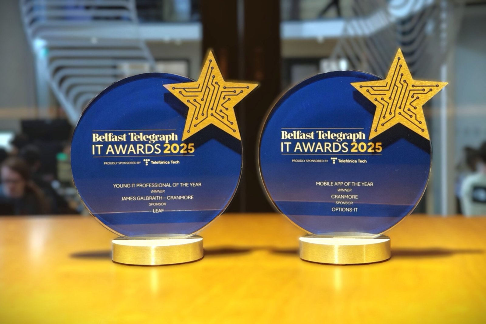 Two blue Belfast Telegraph IT Award trophies on a desk. One for Mobile App of the Year and the other for Young IT Professional of the Year.